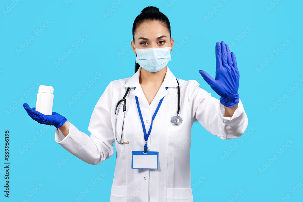 Photo of charming young woman doctor holding bottle of pills and showing stop gesture with hand over blue background.