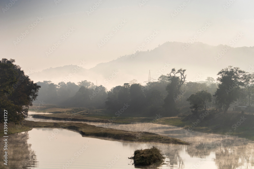 Fototapeta premium A beautiful view of a river with its bank covered with dense evergreen rainforest mountains captured during an early foggy morning at Kaziranga National Park, Assam, Northeast, India.