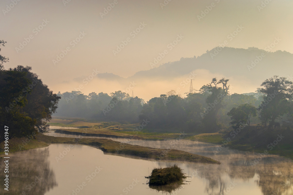 A beautiful view of a river with its bank covered with dense evergreen rainforest mountains captured during an early foggy morning at Kaziranga National Park, Assam, Northeast, India. 