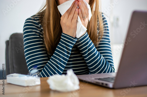Canvas Print Sick woman working on laptop at home, covering nose and mouth when coughing and sneezing in tissue, ill freelance woman with cold