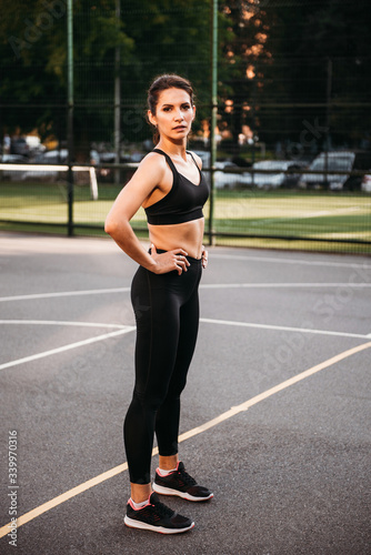 Beautiful sexy fitness trainer wearing black leggings and sport bra standing at the court outside. Energetic fit caucasian dark hair girl posing for the camera. © innatyshchenko