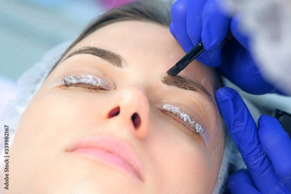 Cosmetologist making lash laminating and eyebrow painting for woman in beauty clinic, face closeup. Beautician applying dark brown paint on woman's brows using brush in cosmetology.