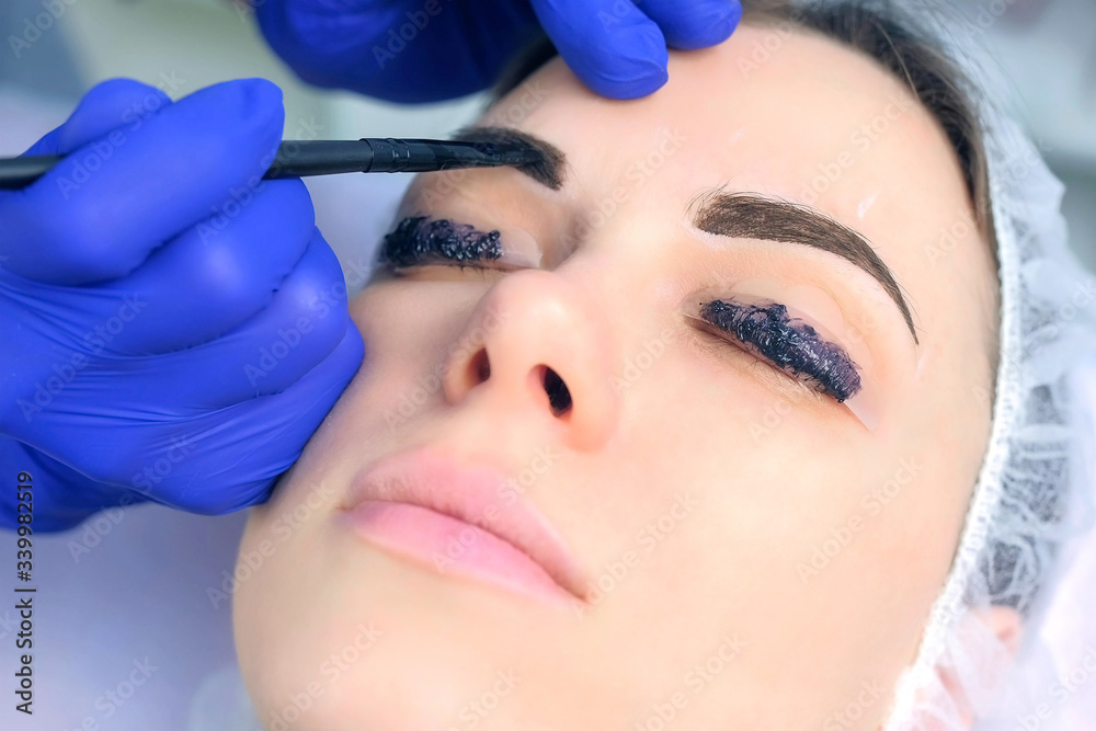 Cosmetologist making lash laminating, eyebrow and lash painting for woman in beauty clinic, face closeup. Beautician applying dark brown paint on woman's brows using brush in cosmetology.