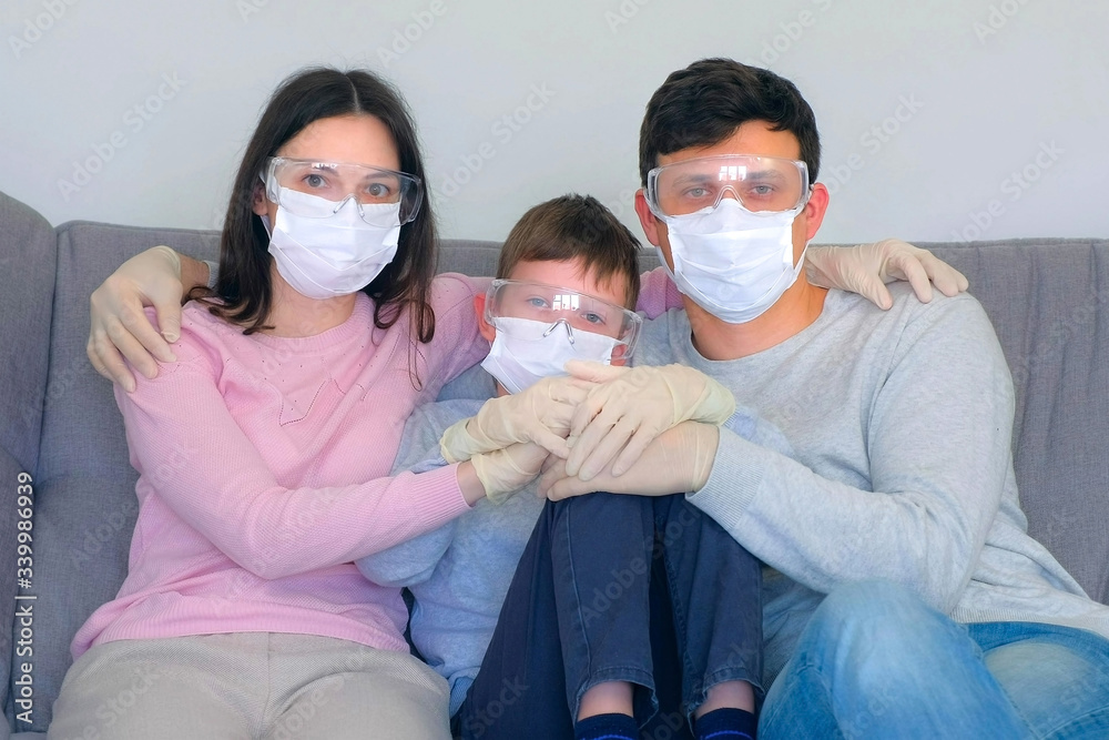 Family mom, dad, son in protective masks, glasses and gloves sitting on couch cuddling at home and looking at camera, coronavirus pandemic quarantine. Worldwide global COVID-19 epidemic. 2019-ncov.