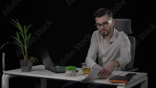 Copywriter reviews article from papers and types on laptop. Bearded young man in eyeglasses at white desk. Indoor studio shot on black background.