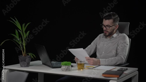 Tired and exhausted copywriter reviews paper sheets with article and throws out bad copies. one by one. Bearded young man in eyeglasses at white desk. Indoor studio shot on black background.