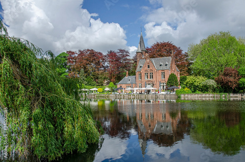 View of tranquil public green space featuring Minnewater Lake and small castle in Bruges during sunny day in spring, Belgium © Maria Vonotna