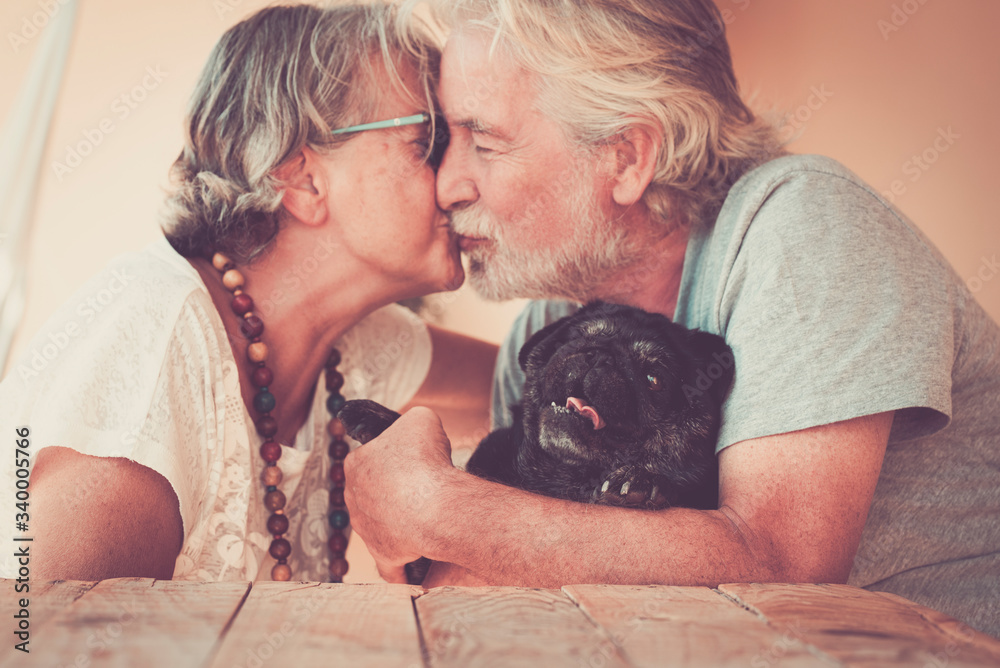 Happy old senior couple people in love kiss eachother and hug their lovely black dog pug - home leisure activity and forever together concept family and relationship Happy old senior couple people in love kiss eachother and hug their lovely black dog pug - home leisure activity and forever together concept family and relationship