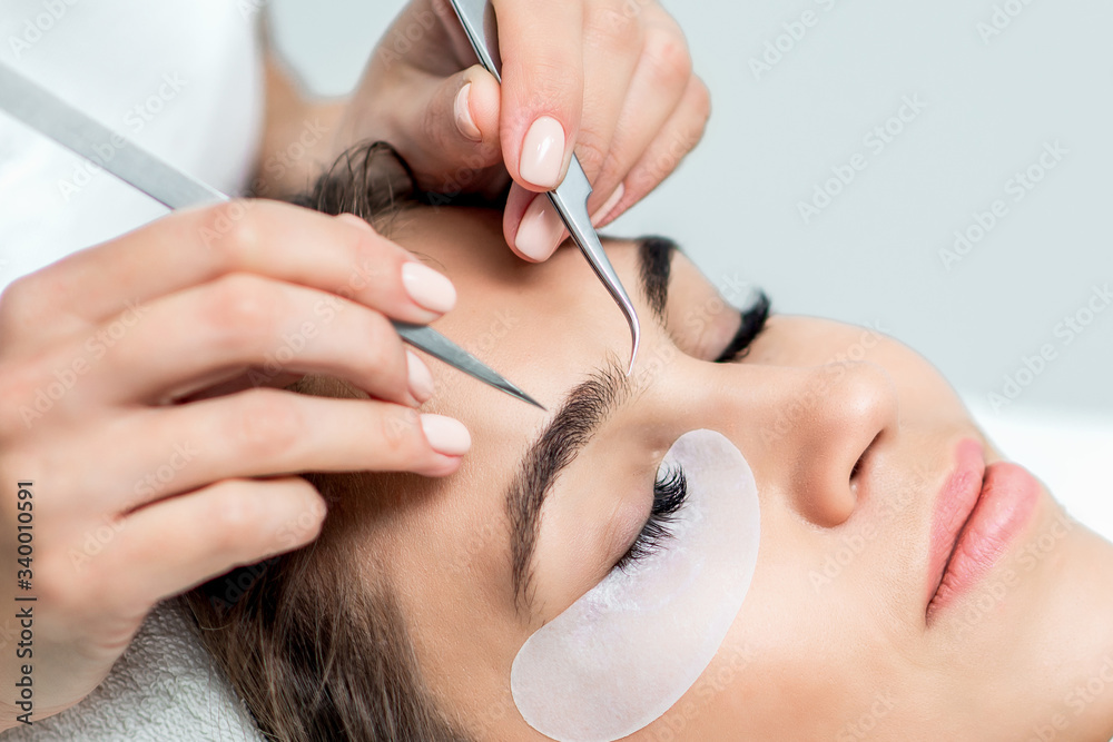 Cosmetologist hands are holding tweezers above the face of young woman during eyelash extensions procedure, close up.