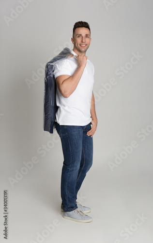 Man holding hanger with jacket in plastic bag on light grey background. Dry-cleaning service
