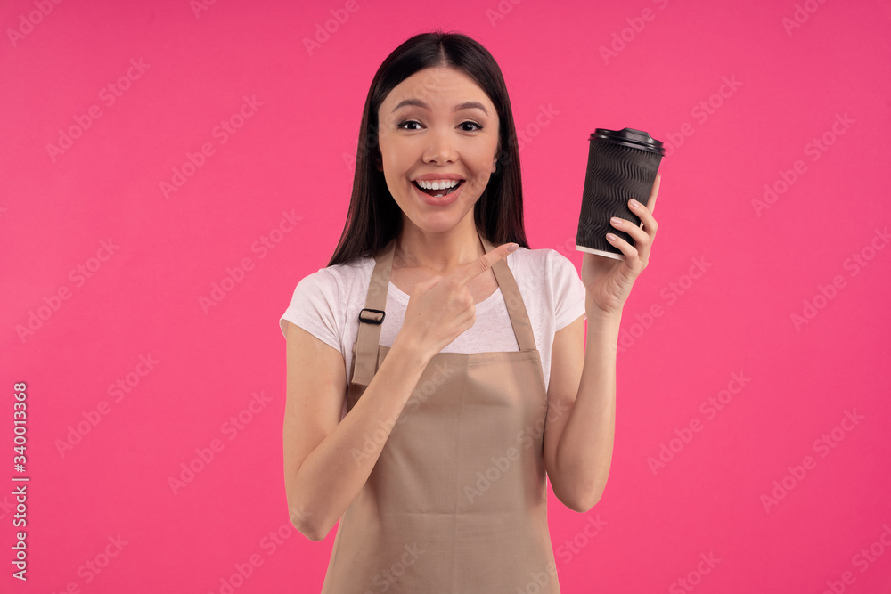 Nice stylish brunette asian girl, barista in casual tshirt and apron, points to a paper cup of tea or coffee, isolated over pink background Nice stylish brunette asian girl, barista in casual tshirt and apron, points to a paper cup of tea or coffee, isolated over pink background