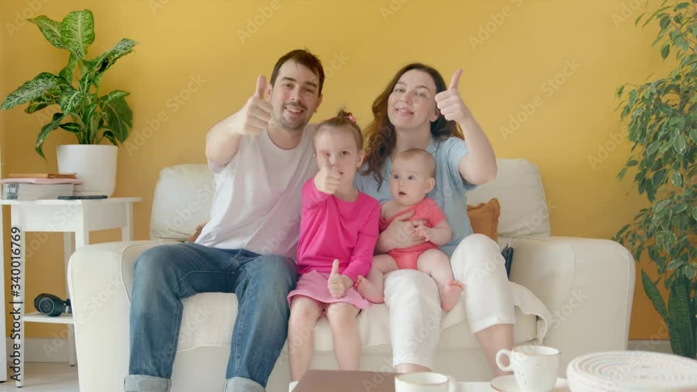 A Young Family, Father, Mother and Two Children Together Give a Thumbs Up. Cozy Interior with Yellow Wall, White Couch and House Plants. A Young Family, Father, Mother and Two Children Together Give a Thumbs Up. Cozy Interior with Yellow Wall, White Couch and House Plants.