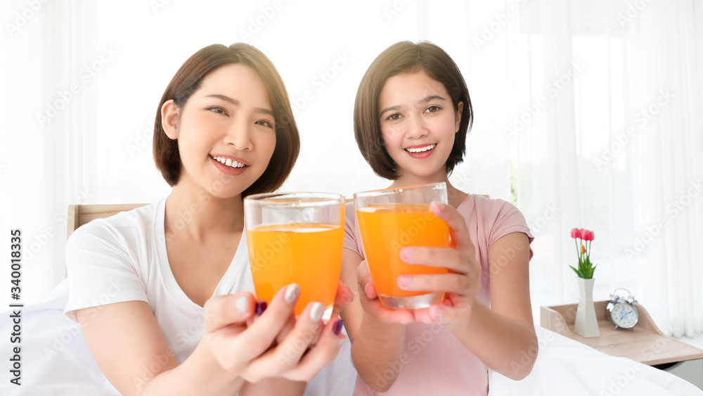 Asian young two woman smile and show closeup glass of organic orange juice beverage with nutrition and vitamin C for breakfast fresh drink within sunlight in the morning from bedroom at home Asian young two woman smile and show closeup glass of organic orange juice beverage with nutrition and vitamin C for breakfast fresh drink within sunlight in the morning from bedroom at home