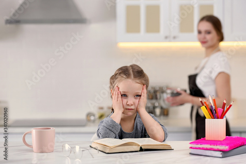 Tired little girl doing homework while mother is busy in kitchen