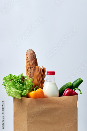 Groceries: bread, buckwheat spaghetti, milk, lettuce, two bell peppers and two cucumbers all in one paper shopping bag on white background