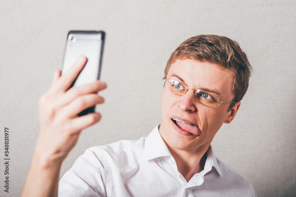The man in glasses makes a Selfe jokingly phone. On a gray background. The man in glasses makes a Selfe jokingly phone. On a gray background.