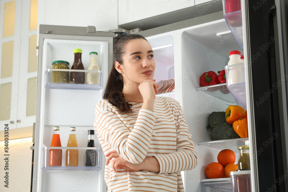 Thoughtful young woman near open refrigerator indoors