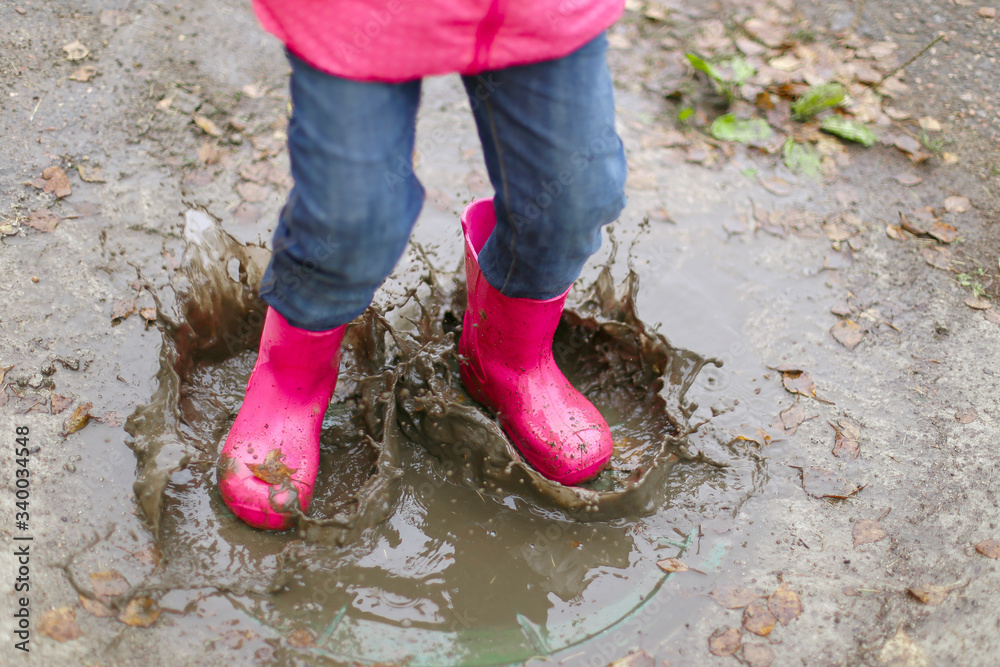 Happy little girl in pink waterproof jacket, rubber boots cheerfully jumps through puddles on street road in rainy weather. Spring, autumn. Children's fun in fresh air after rain. Outdoors recreation