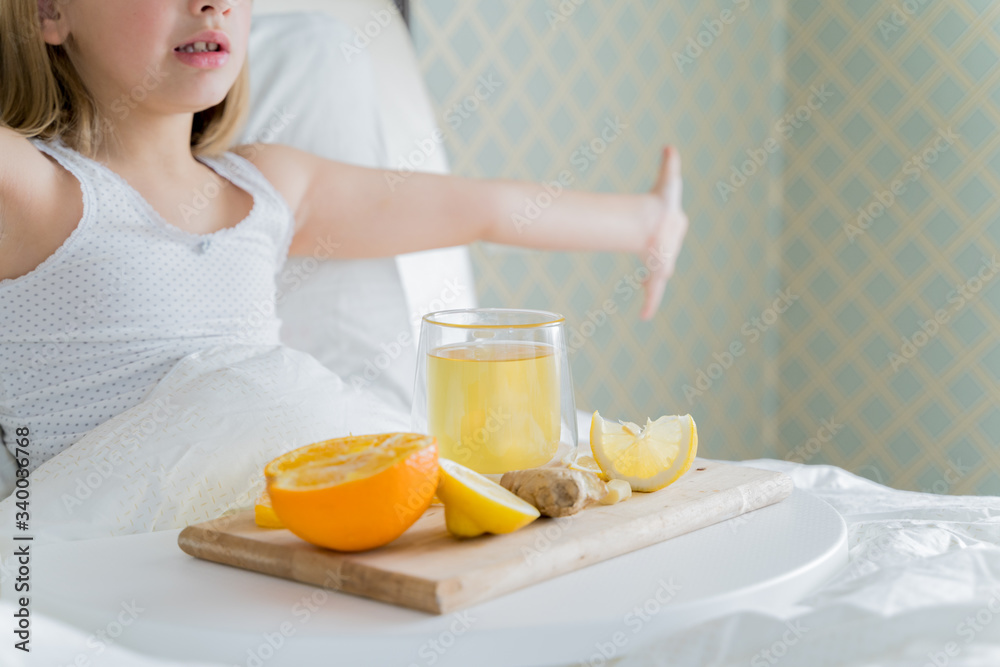 Little sick girl in bed with cup of antipyretic drugs for colds,flu.Tea with citrus vitamin C,ginger root,lemon,orange.Wooden tray. Home self-treatment.Medical quarantine covid-19 coronavirus therapy