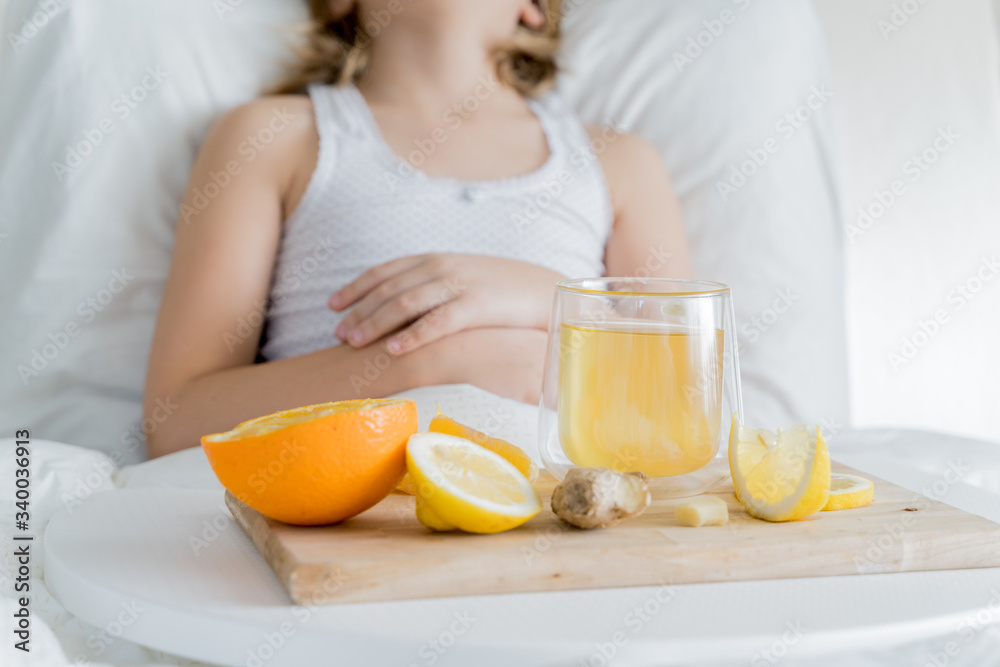 Little sick girl in bed with cup of antipyretic drugs for colds,flu.Tea with citrus vitamin C,ginger root,lemon,orange.Wooden tray. Home self-treatment.Medical quarantine covid-19 coronavirus therapy
