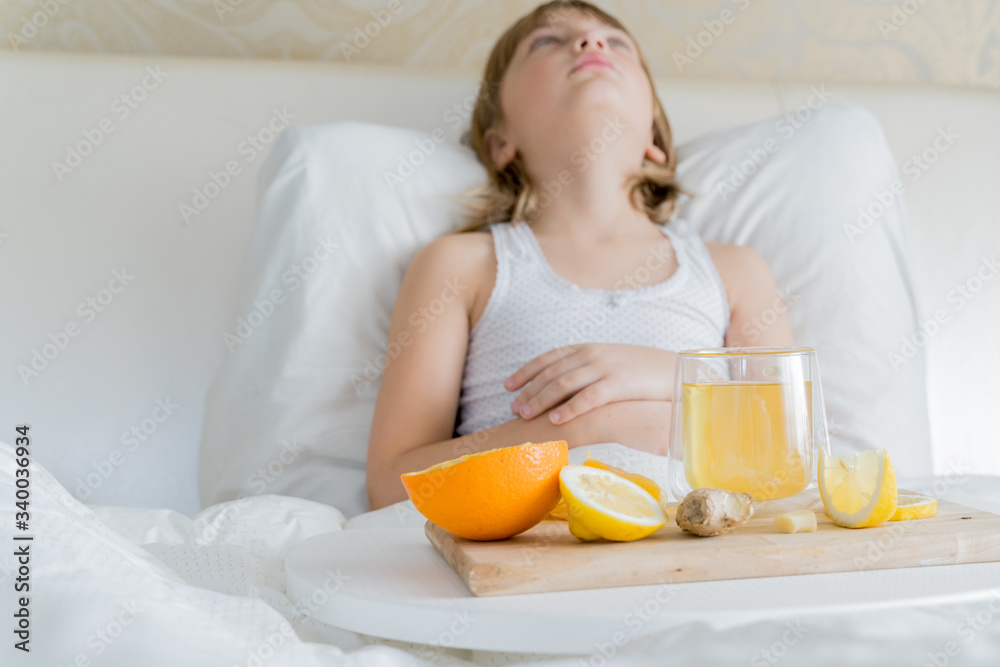 Little sick girl in bed with cup of antipyretic drugs for colds,flu.Tea with citrus vitamin C,ginger root,lemon,orange.Wooden tray. Home self-treatment.Medical quarantine covid-19 coronavirus therapy
