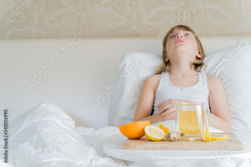 Little sick girl in bed with cup of antipyretic drugs for colds,flu.Tea with citrus vitamin C,ginger root,lemon,orange.Wooden tray. Home self-treatment.Medical quarantine covid-19 coronavirus therapy