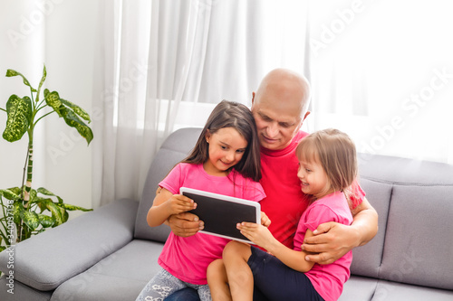 An elderly man and a little girl sitting on his lap are sitting in a room. Grandfather holding a tablet in his hand and looking at granddaughter