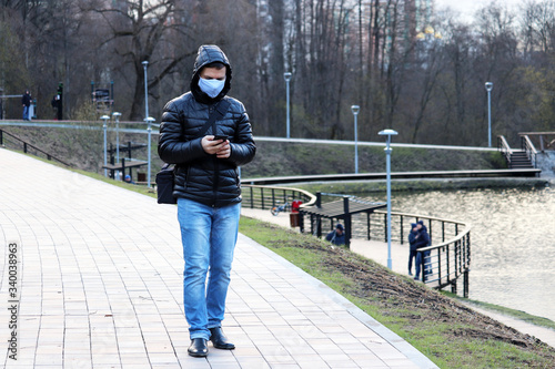 Coronavirus protection, man in medical mask walking on a city park with smartphone in hands. Concept of quarantine during covid-19 pandemic