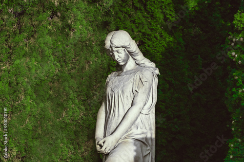 sculpture of saint woman against green thuja in old cemetery. stoned angel with closed eyes monument at cemetery. Graveyard old grey statue on funeral. Death, loss empty natural green background.