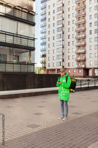 Young male food courier in green uniform using navigation app or texting on cell phone while standing in city street with insulated backpack delivering order © Comeback Images