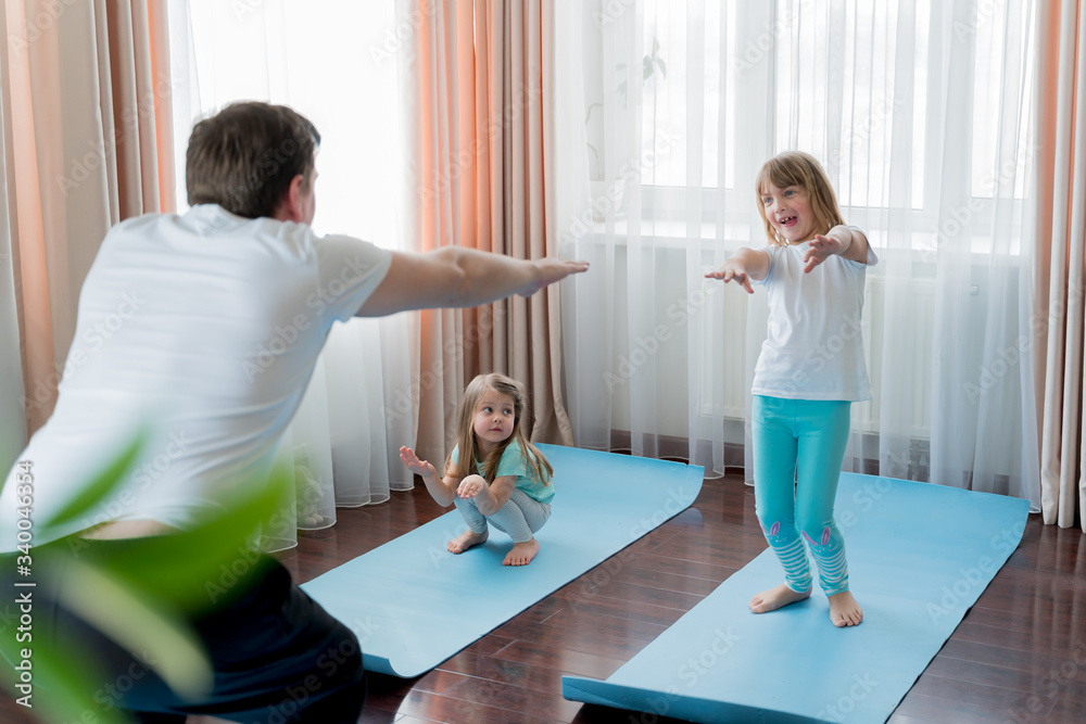 Fototapeta premium Home workout in flat.Dad teaching little girls fitness, daughters. Physical morning exercises on blue yoga mats.Family children fun.Healthy indoor activity.Quarantine,coronavirus covid-19.No equipment