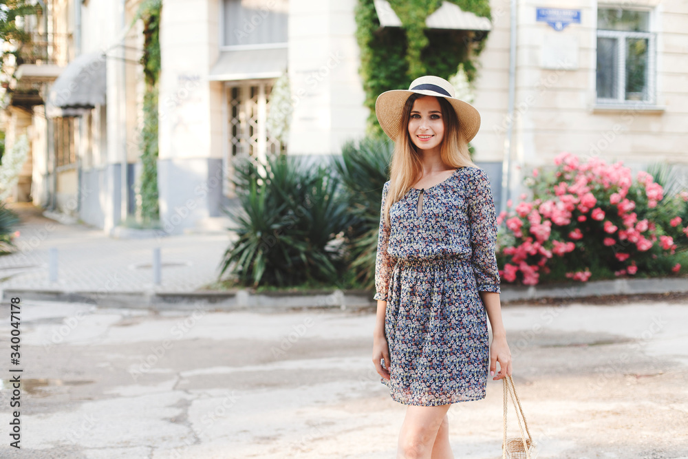 Young beautiful woman with long hair, dressed in a light blue dress with a floral print and straw boater hat, walks  in the summer on the city street near the flowering pink rose bushes.