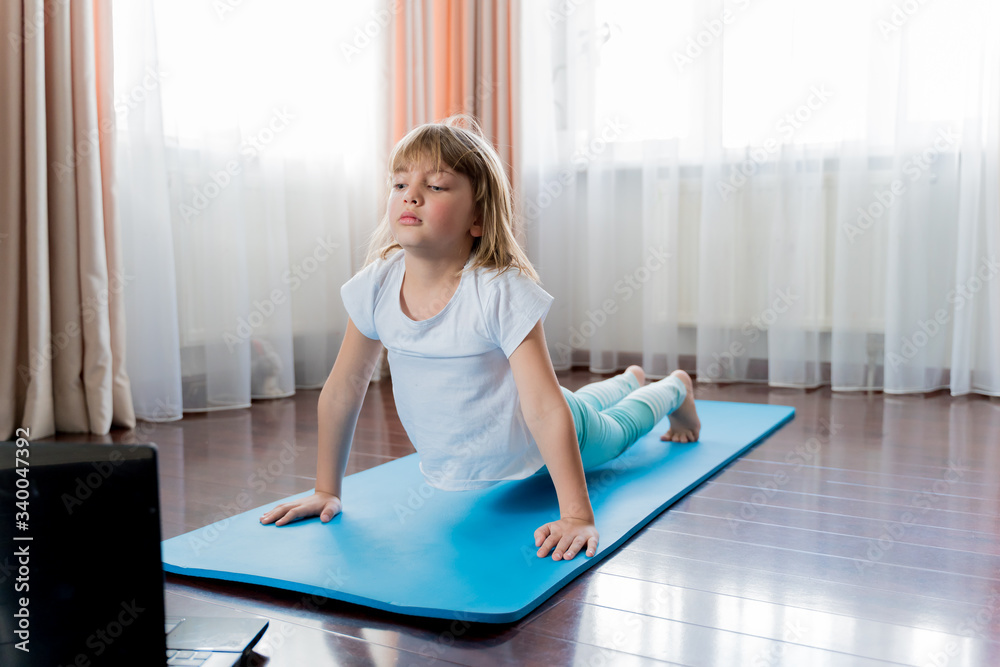 Little girl make home online laptop workout in flat.Baby yoga,fitness training.Distance school physical morning exercises on blue mat.Healthy activity,fun.Quarantine,coronavirus,covid-19.No equipment Little girl make home online laptop workout in flat.Baby yoga,fitness training.Distance school physical morning exercises on blue mat.Healthy activity,fun.Quarantine,coronavirus,covid-19.No equipment