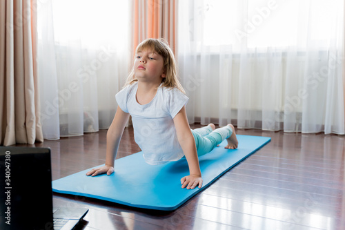 Little girl make home online laptop workout in flat.Baby yoga,fitness training.Distance school physical morning exercises on blue mat.Healthy activity,fun.Quarantine,coronavirus,covid-19.No equipment