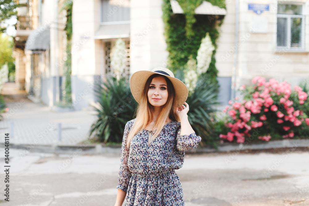 Young beautiful woman with long hair, dressed in a light blue dress with a floral print and straw boater hat, walks  in the summer on the city street near the flowering pink rose bushes.