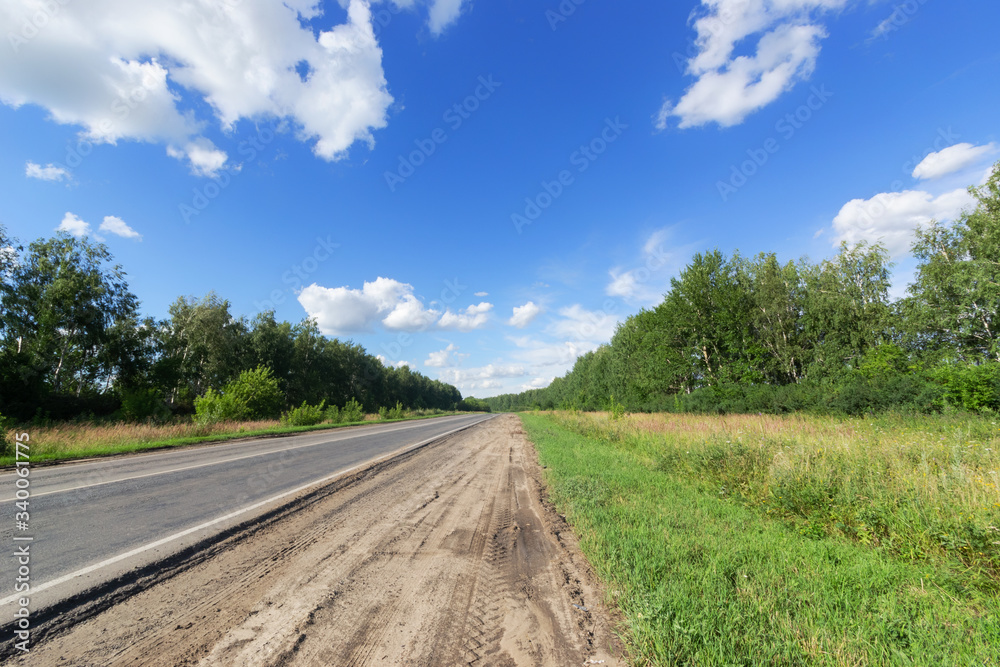 Paved and unpaved road, among birch trees under a blue sky and white clouds