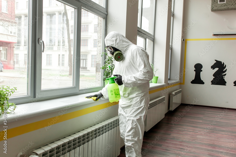 A man in protective equipment disinfects with a sprayer in the office. Surface treatment due to coronavirus covid-19 disease. A man in a white suit disinfects the room with a spray gun. Virus pandemic A man in protective equipment disinfects with a sprayer in the office. Surface treatment due to coronavirus covid-19 disease. A man in a white suit disinfects the room with a spray gun. Virus pandemic