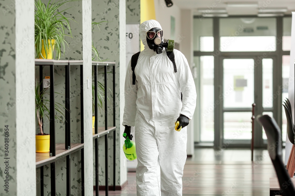 A man in protective equipment disinfects with a sprayer in the office. Surface treatment due to coronavirus covid-19 disease. A man in a white suit disinfects the room with a spray gun. Virus pandemic