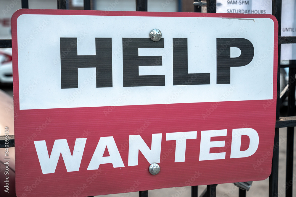 A red black and white sign with help wanted text hanging on a black cast iron fence along the sidewalk with two bolts in Chicago during the COVID-19 outbreak and pandemic. A red black and white sign with help wanted text hanging on a black cast iron fence along the sidewalk with two bolts in Chicago during the COVID-19 outbreak and pandemic.