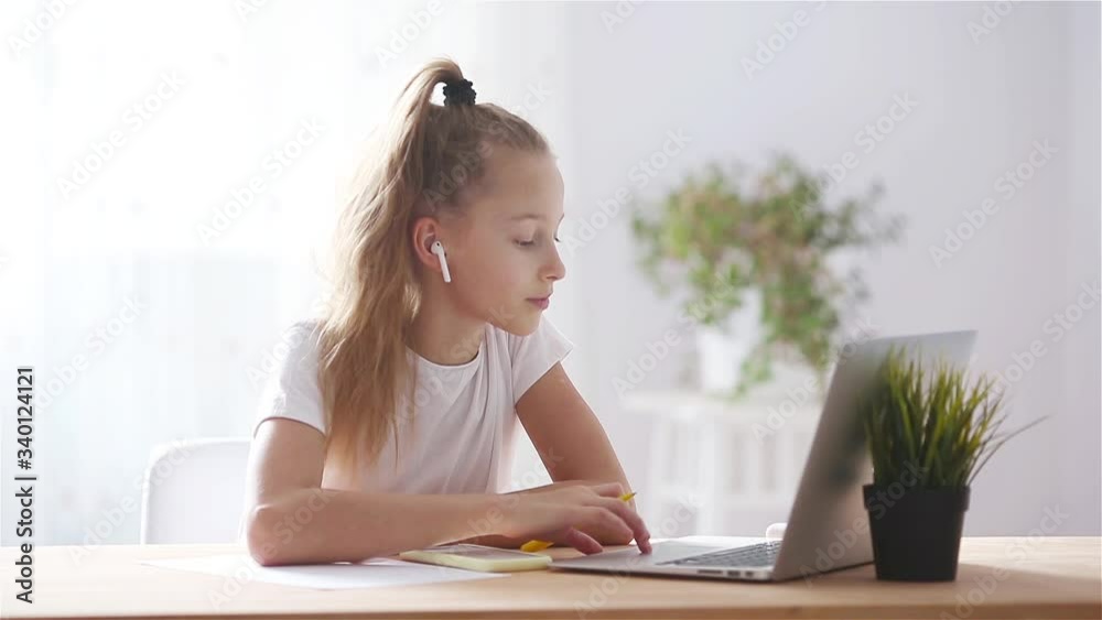 Serious schoolgirl sitting at table with laptop and textbook and doing homework. Serious schoolgirl sitting at table with laptop and textbook and doing homework.