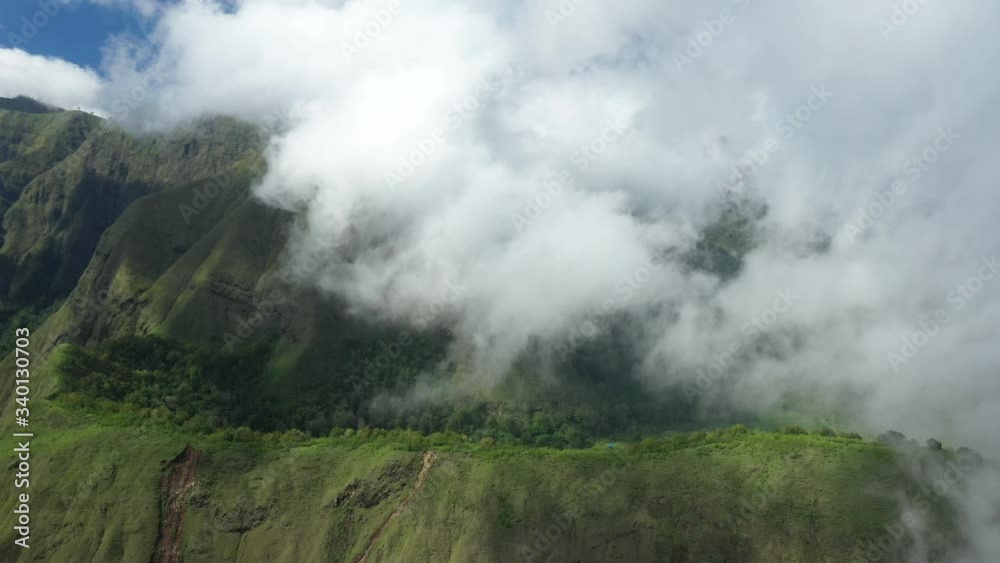 View from above, stunning aerial view of a green mountain range surrounded by clouds and a beautiful blue sky in the background. Sembalun, Lombok, West Nusa Tenggara, Indonesia. View from above, stunning aerial view of a green mountain range surrounded by clouds and a beautiful blue sky in the background. Sembalun, Lombok, West Nusa Tenggara, Indonesia.