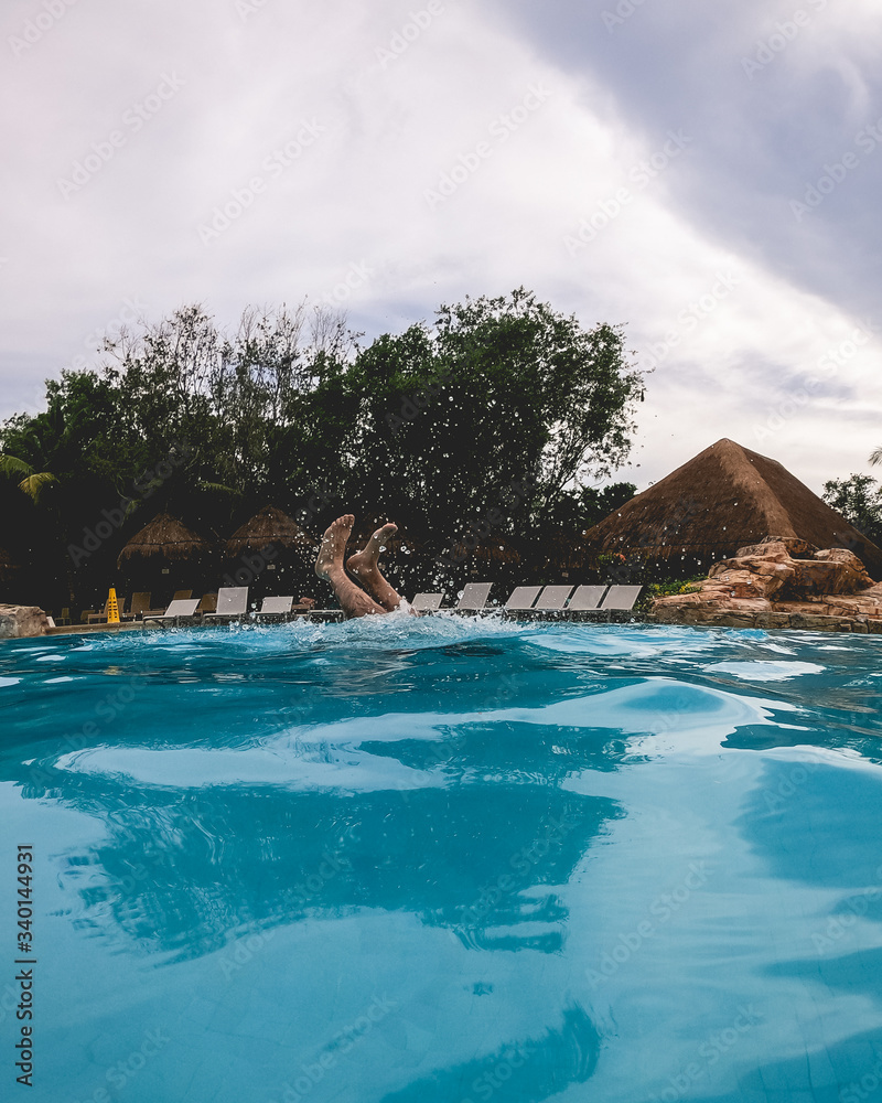 Feet of man at beautiful pool with buildings with straw roof, trees, and cloudy sky, Mexico
