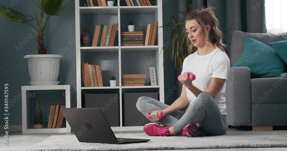 oung sporty woman in activewear sitting with crossed legs on floor during video call on laptop with fitness instructor. Beautiful girl training with dumbbells at home. oung sporty woman in activewear sitting with crossed legs on floor during video call on laptop with fitness instructor. Beautiful girl training with dumbbells at home.