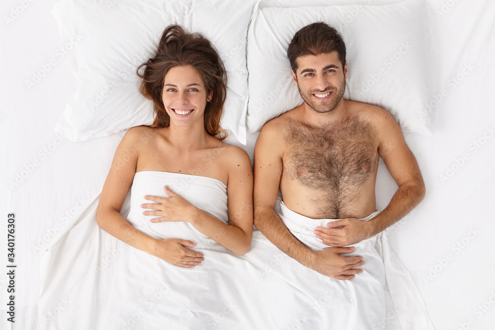 Top view of happy spouses awake in one bed, enjoy good morning, look directly at camera with smile, feel comfort and relaxed, have intimacy and closeness, spend free time at home. Horizontal photo