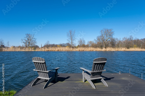 Rustic hand crafted chairs on a wooden deck overlooking a river or lake with reeds in spring sunshine