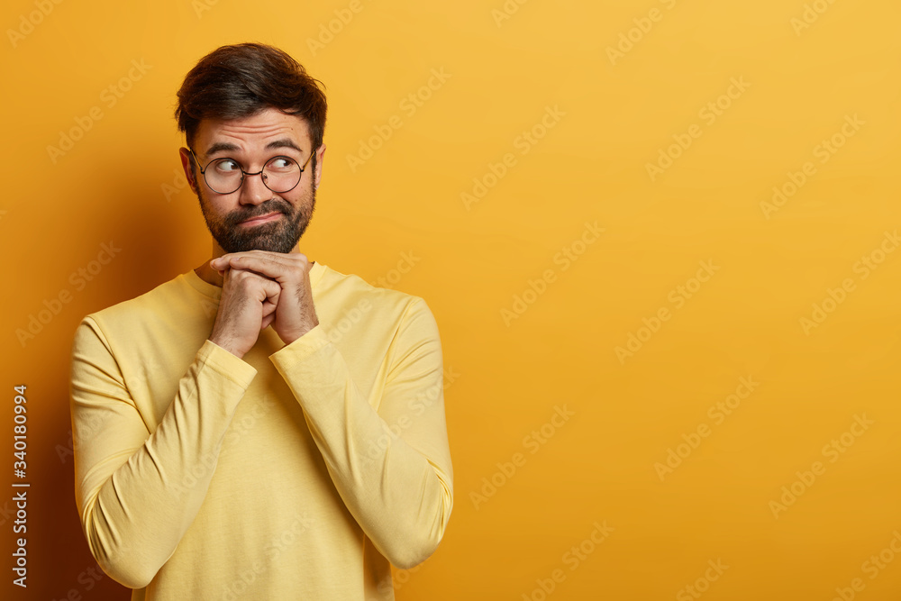 Waist up shot of bearded young man thinks seriously about your offer, keeps hands under chin, concentrated aside, wears optical glasses and sweater, dreams about something, isolated on yellow wall Waist up shot of bearded young man thinks seriously about your offer, keeps hands under chin, concentrated aside, wears optical glasses and sweater, dreams about something, isolated on yellow wall