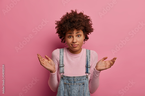 Unsure questioned dark skinned woman raises hands up with confused expression, puzzled to give answer on difficult question, wears casual outfit, isolated over pink background. Human perception