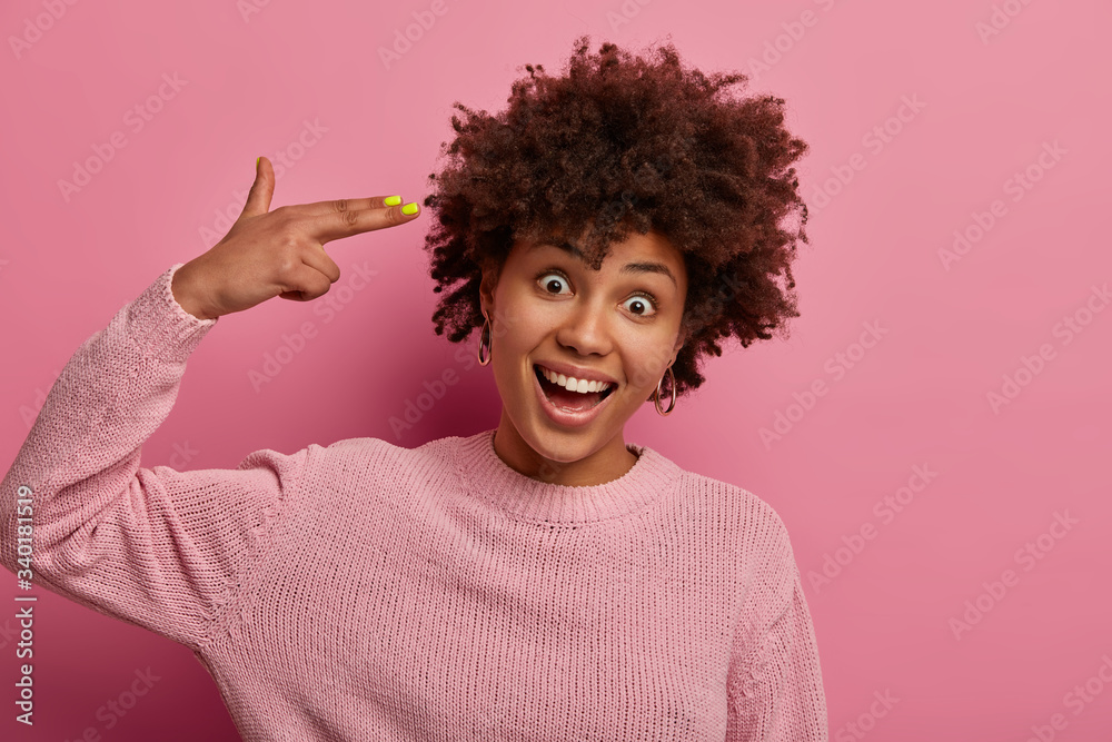 Happy playful African American woman makes finger gun pistol, laughs positively, tilts head, wears rosy jumper, has positive mood, smiles broadly, isolated on pink background. Body language concept Happy playful African American woman makes finger gun pistol, laughs positively, tilts head, wears rosy jumper, has positive mood, smiles broadly, isolated on pink background. Body language concept