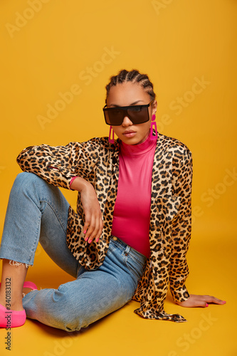 Photo of dark skinned lady in trendy sunglasses and leopard shirt, poses on floor against yellow background, has own style, cornrow hairstyle. Vertical shot of stylish woman in stylish clothes