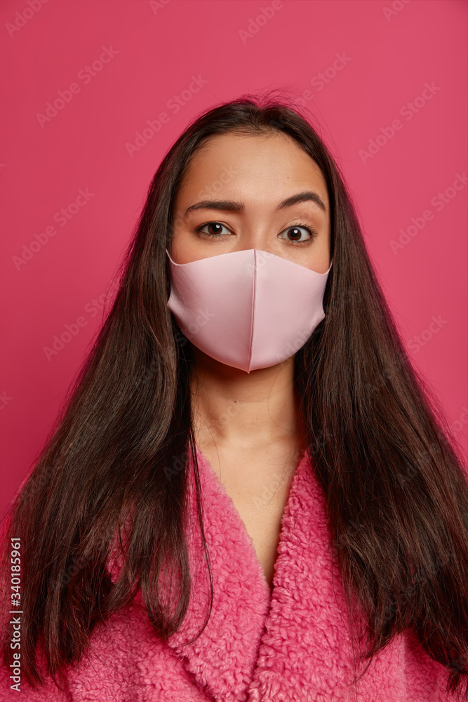 Stay at home. Photo of brunette woman wears handmade protective mask from virus, soft bath robe, being on self isolation during quarantine, stands alone against pink background. Health care. Stay at home. Photo of brunette woman wears handmade protective mask from virus, soft bath robe, being on self isolation during quarantine, stands alone against pink background. Health care.
